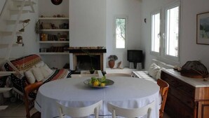 A cozy living room with a fireplace, a round dining table with chairs, a bookshelf, and a sofa. - Eretria: Single storey house, in an olive grove, 250 from the beach (Eretria)