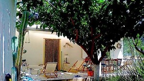 An outdoor patio area with a table and chairs, a small stage, and a tree providing shade. - Eretria: Single storey house, in an olive grove, 250 from the beach (Eretria)