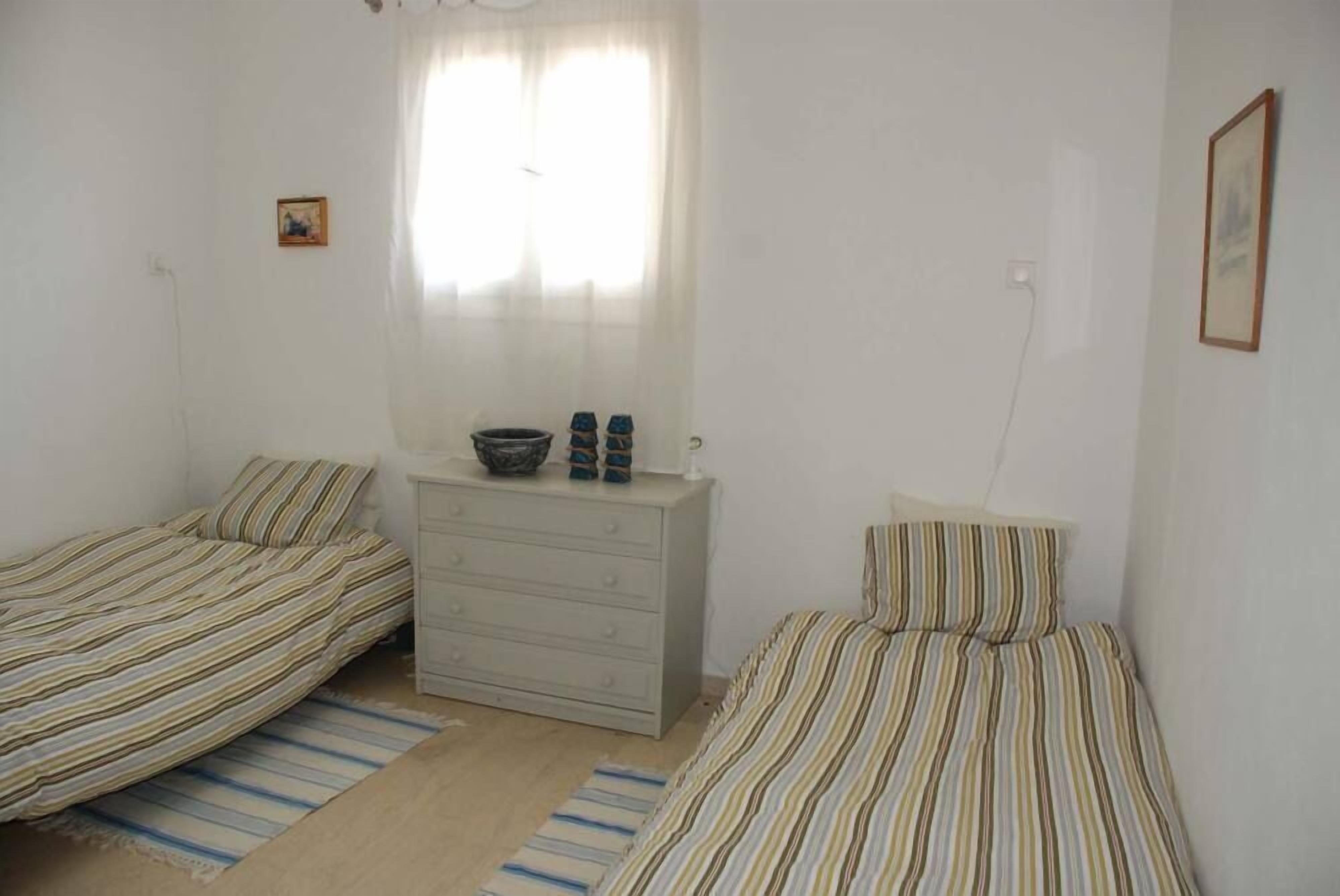 Une petite chambre propre, avec deux lits simples, une commode et une fenêtre ornée de rideaux légers.