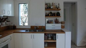 A kitchen with white cabinets, a wooden countertop, a built-in oven, a sink, and shelves with kitchen items. - Eretria: Single storey house, in an olive grove, 250 from the beach (Eretria)
