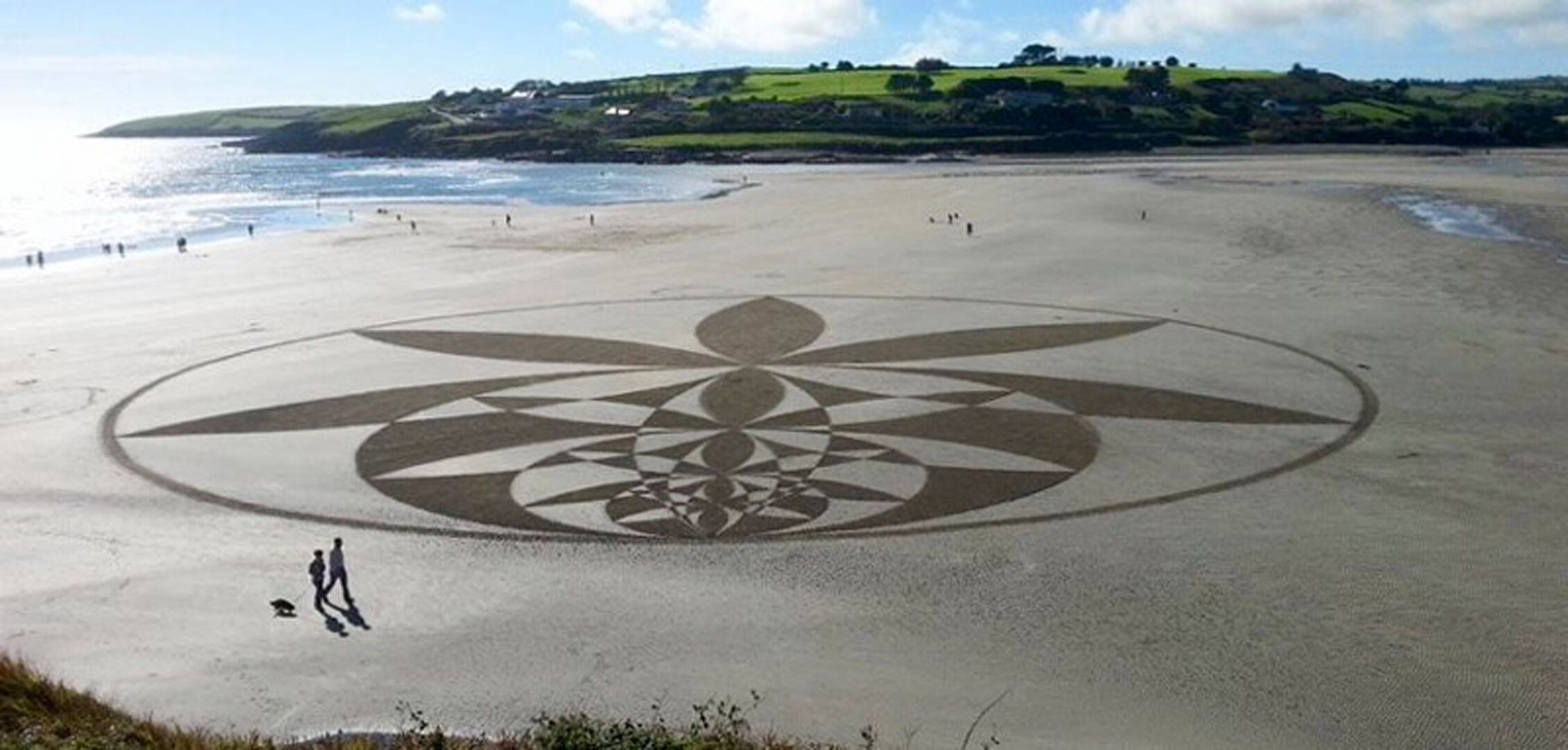 A beach with a large geometric pattern in the sand, people walking, and a hilly landscape in the background.