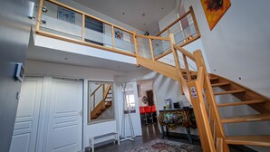 Een ruime kamer met een houten trap naar een hoger niveau, een grote spiegel en een eethoek met rode stoelen.