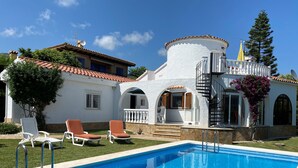 Un espace aménagé au bord d’une piscine à l’eau d’un bleu limpide, avec des murs blancs et un toit recouvert de tuiles rouges. On y trouve des chaises longues et un escalier menant à un balcon.
