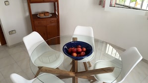 Une table ronde en verre, entourée de chaises blanches, un saladier bleu rempli de fruits et un buffet en bois sur lequel sont posées des tasses.