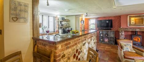 A cozy kitchen with a brick island, wooden chairs, a TV, and a fireplace.