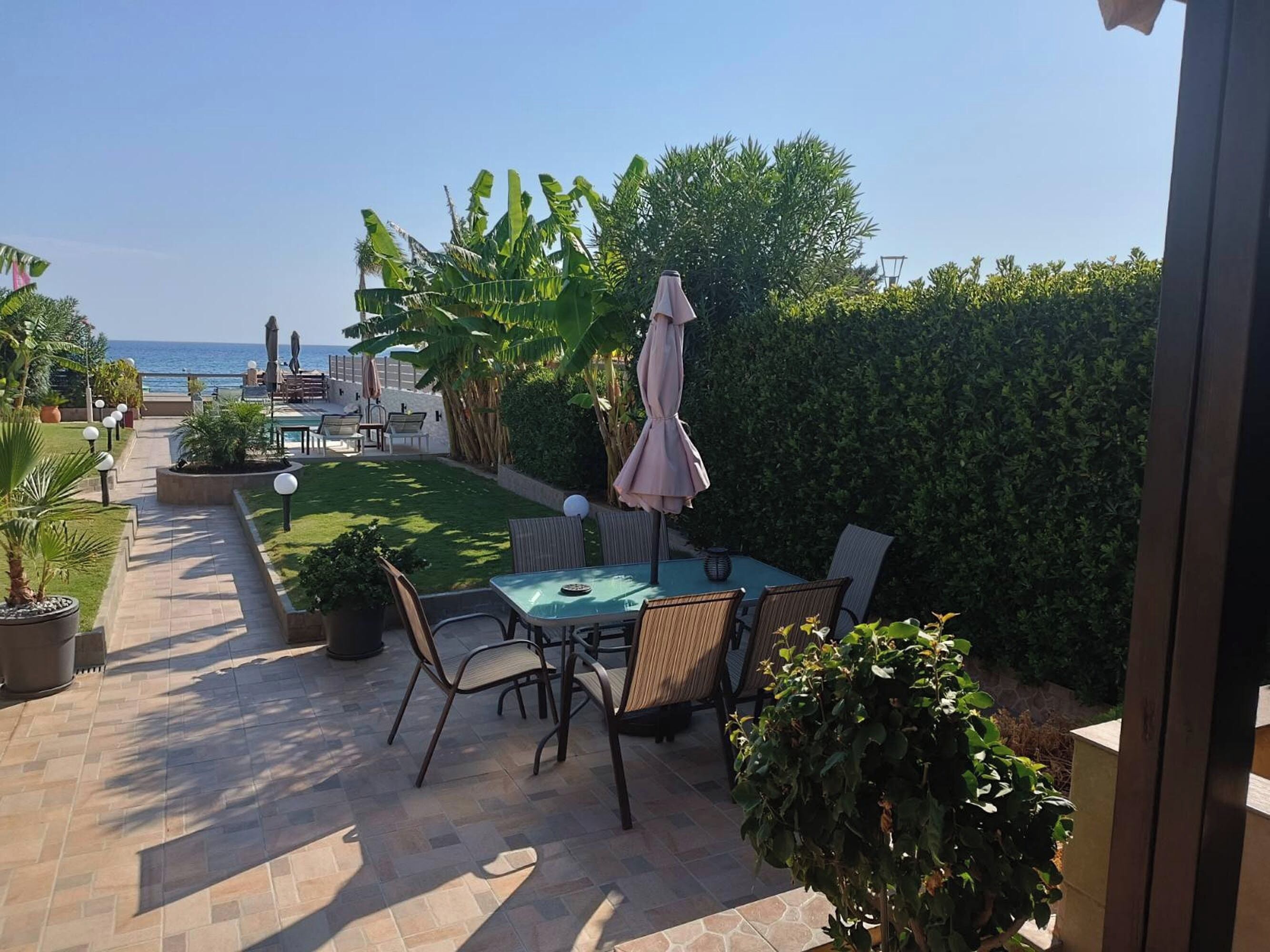 Un patio con mesa y sillas, vista al mar y un jardín cuidado con diversas plantas.