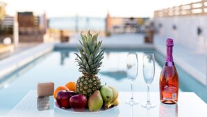Een tafel met een fles rosé wijn, twee glazen, een fruitschaal en een zeepstaafje bij een zwembad.