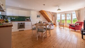 Une cuisine spacieuse avec des meubles blancs, un coin repas avec une table et des chaises en bois, et un fauteuil rouge près d’une grande fenêtre.