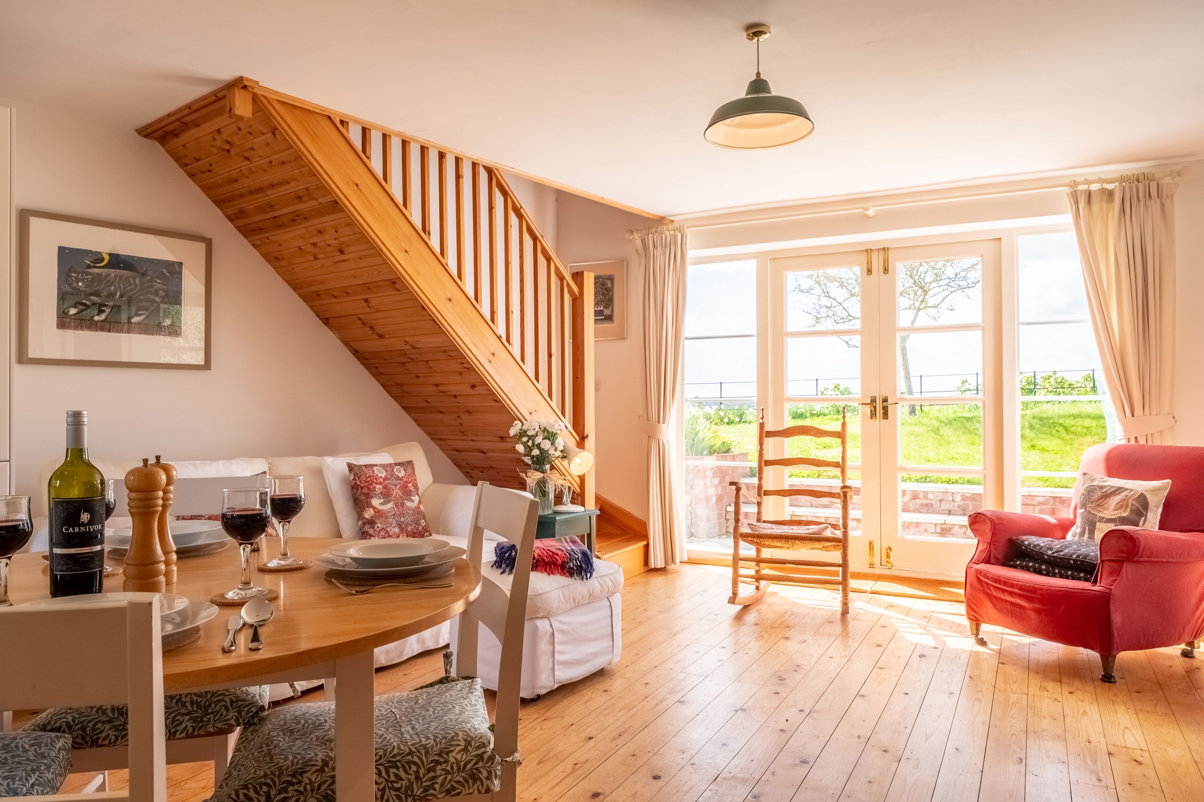 A cozy living room with a wooden staircase, a dining table set with wine glasses, and a red armchair.