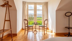 Une pièce avec un plancher en bois, une table entourée de chaises, un portant suspendu et une vue sur un paysage verdoyant par la fenêtre.