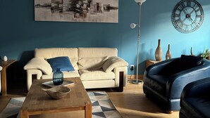 Una sala de estar con un sofá color beige, un sillón azul, una mesa de centro de madera, un tapete geométrico, un reloj de pared y un cuadro.