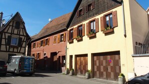 Vista de calle con arquitectura europea tradicional, incluyendo un edificio con entramado de madera y un edificio moderno con contraventanas de madera y plantas en macetas.