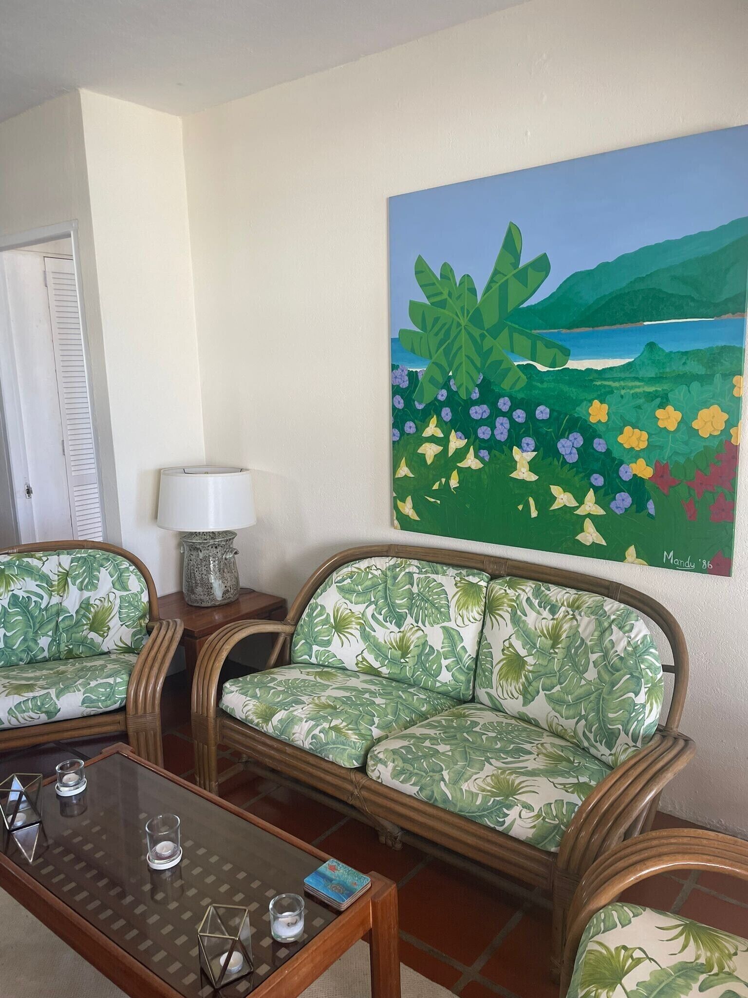 A room with a sofa and chairs with a tropical leaf pattern, a glass coffee table, and a painting of a landscape with mountains and flowers.
