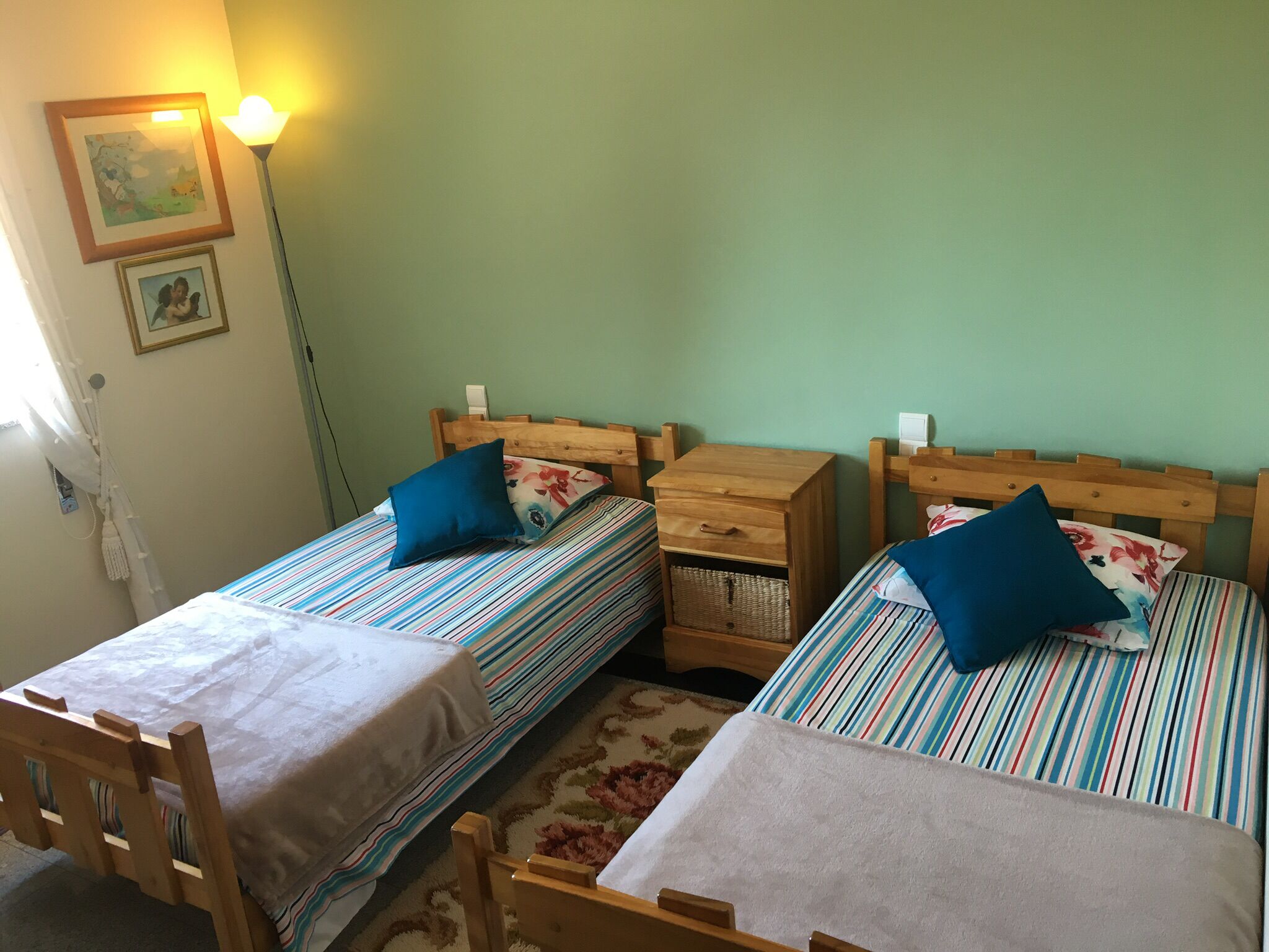 Een kamer met twee eenpersoonsbedden, elk met een houten hoofdbord en een gestreept beddegoed. Daartussen staat een nachtkastje met een lade, en aan de muur is een lamp te zien.