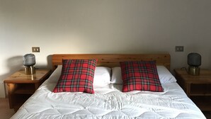 Et soveværelse med sengegavl i træ, hvidt sengetøj og to ternede puder. Der er to natborde med lamper, og et billede hænger i en ramme på væggen.
