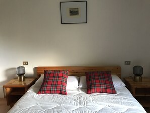 Une chambre à coucher avec une tête de lit en bois, une literie blanche et deux coussins à carreaux. On y trouve deux tables de chevet avec des lampes et un tableau encadré accroché au mur.