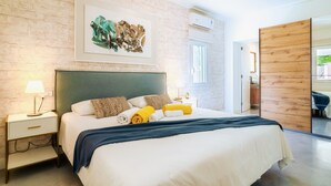 Una habitación de hotel moderna con pared de ladrillo, una cama grande con ropa blanca, cabecera de madera y una obra de arte enmarcada sobre la cama.