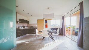 Et moderne køkken med mørke skabe, et spiseområde med hvide stole og en stor skydedør, der fører ud til en balkon med krukkeplanter.