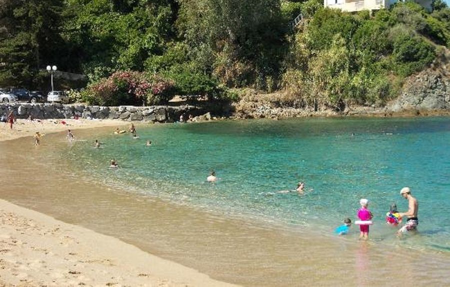 Una playa con agua turquesa cristalina, gente nadando y jugando, y una casa en una colina al fondo.