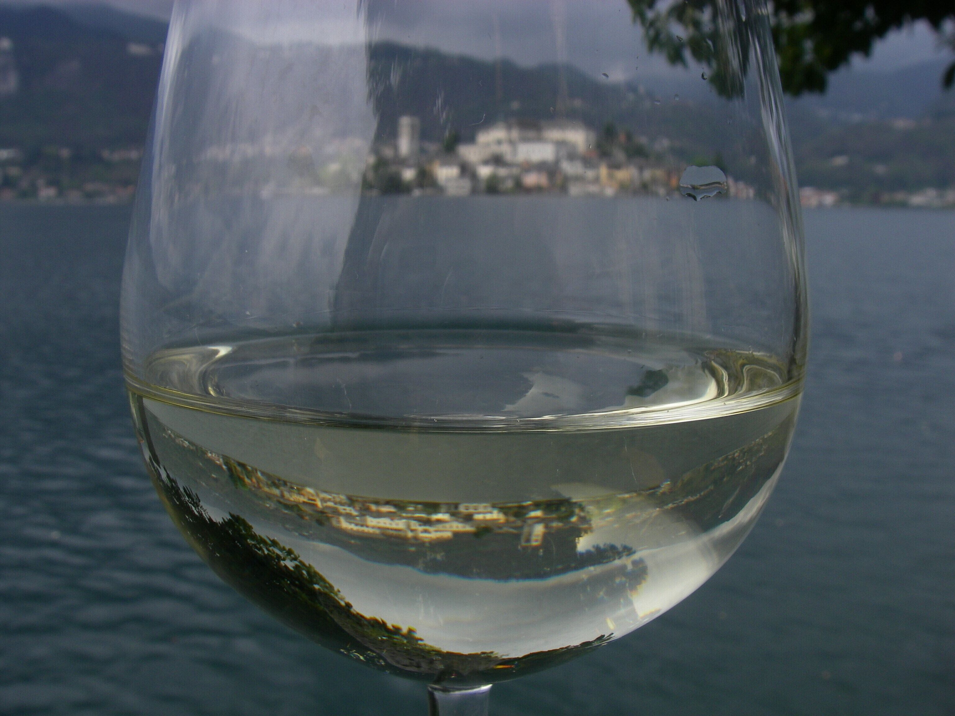 Un verre transparent rempli de vin blanc, dans lequel se reflète un paysage montagneux et des bâtiments au loin.