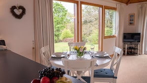 A dining area with a table set for four, a bowl of fruit on the table, and a view of the outdoors through large windows. - Comfortable Cottage With Spectacular Sea And Mountain Views (Duisdale)