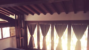A room with a bed, wooden beams on the ceiling, and multiple windows with curtains.