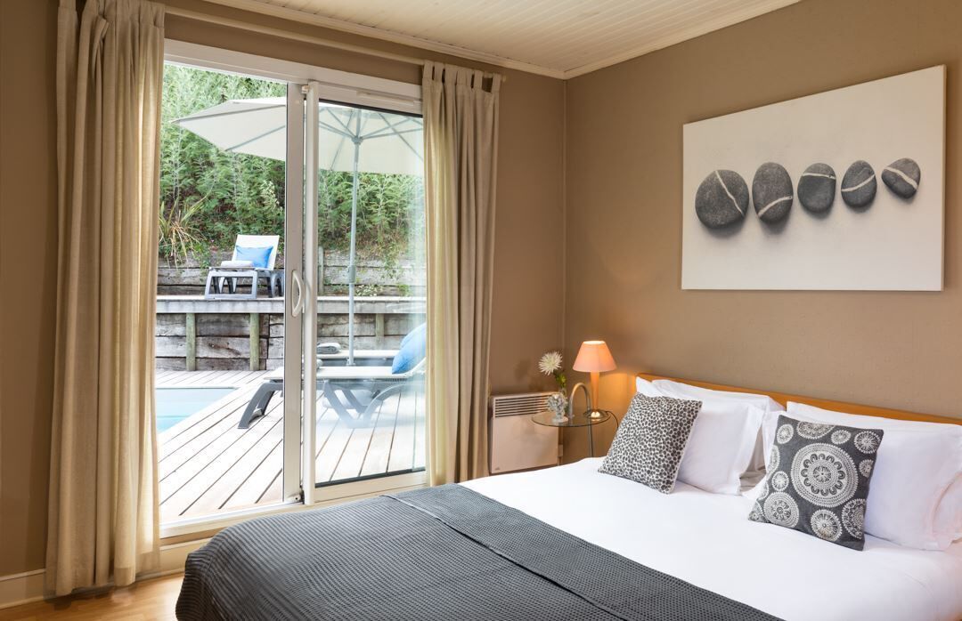 Ein Hotelzimmer mit Bett, Nachttisch mit Lampe, Fenster mit Vorhängen und Blick auf einen Außenpoolbereich.