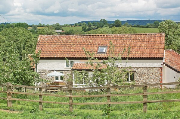 A rustic stone house with a red-tiled roof, surrounded by lush greenery and a wooden fence.
