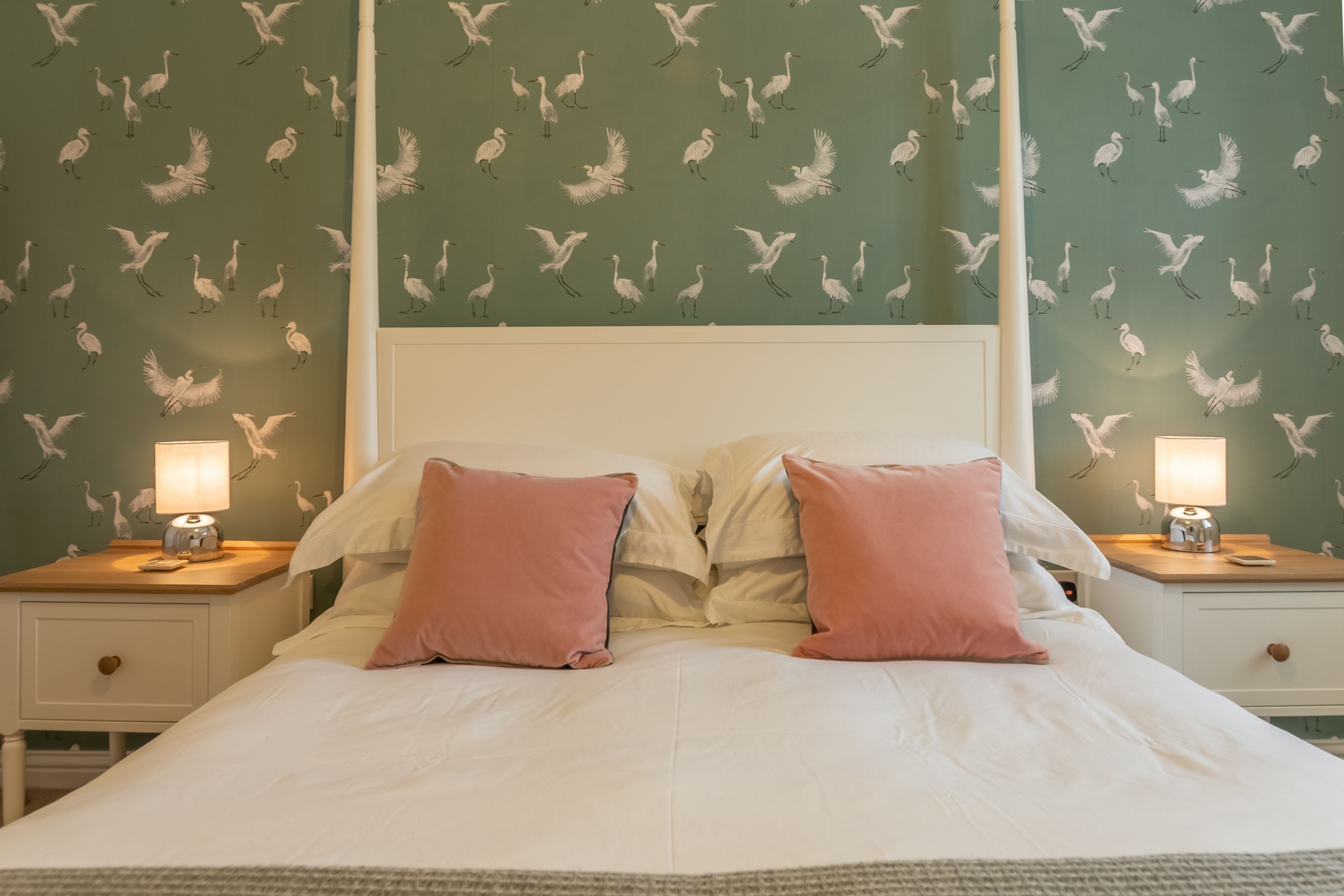 A neatly made bed with white linens and pink pillows, flanked by two bedside tables with lamps, set against a wall with a bird pattern wallpaper.
