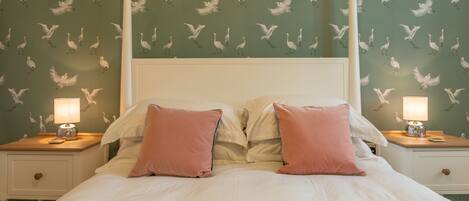 A neatly made bed with white linens and pink pillows, flanked by two bedside tables with lamps, set against a wall with a bird pattern wallpaper.