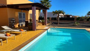 A swimming pool with lounge chairs and a pergola, surrounded by palm trees and a building with a tiled patio. - Luxury 3Bed Villa, Large Heated Pool, Wifi, Sun Terrace, Sea & Golf Course Views (Caleta De Fuste)