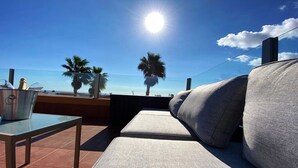 A sunlit outdoor seating area with a view of the sea, featuring a cushioned sofa, a small table, and a metal bucket. - Luxury 3Bed Villa, Large Heated Pool, Wifi, Sun Terrace, Sea & Golf Course Views (Caleta De Fuste)