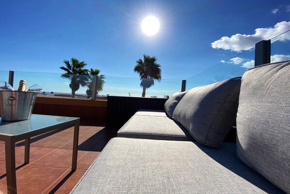Zona de estar al aire libre con luz solar y vistas al mar, con un sofá acolchado, una mesita y un cubo de metal.