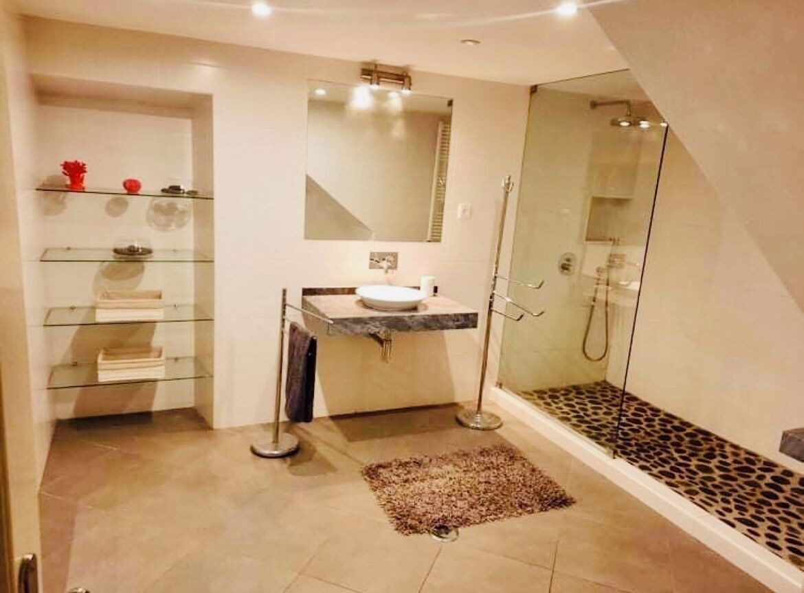 A modern bathroom with a glass shower enclosure, a marble sink, and a glass shelving unit with toiletries.