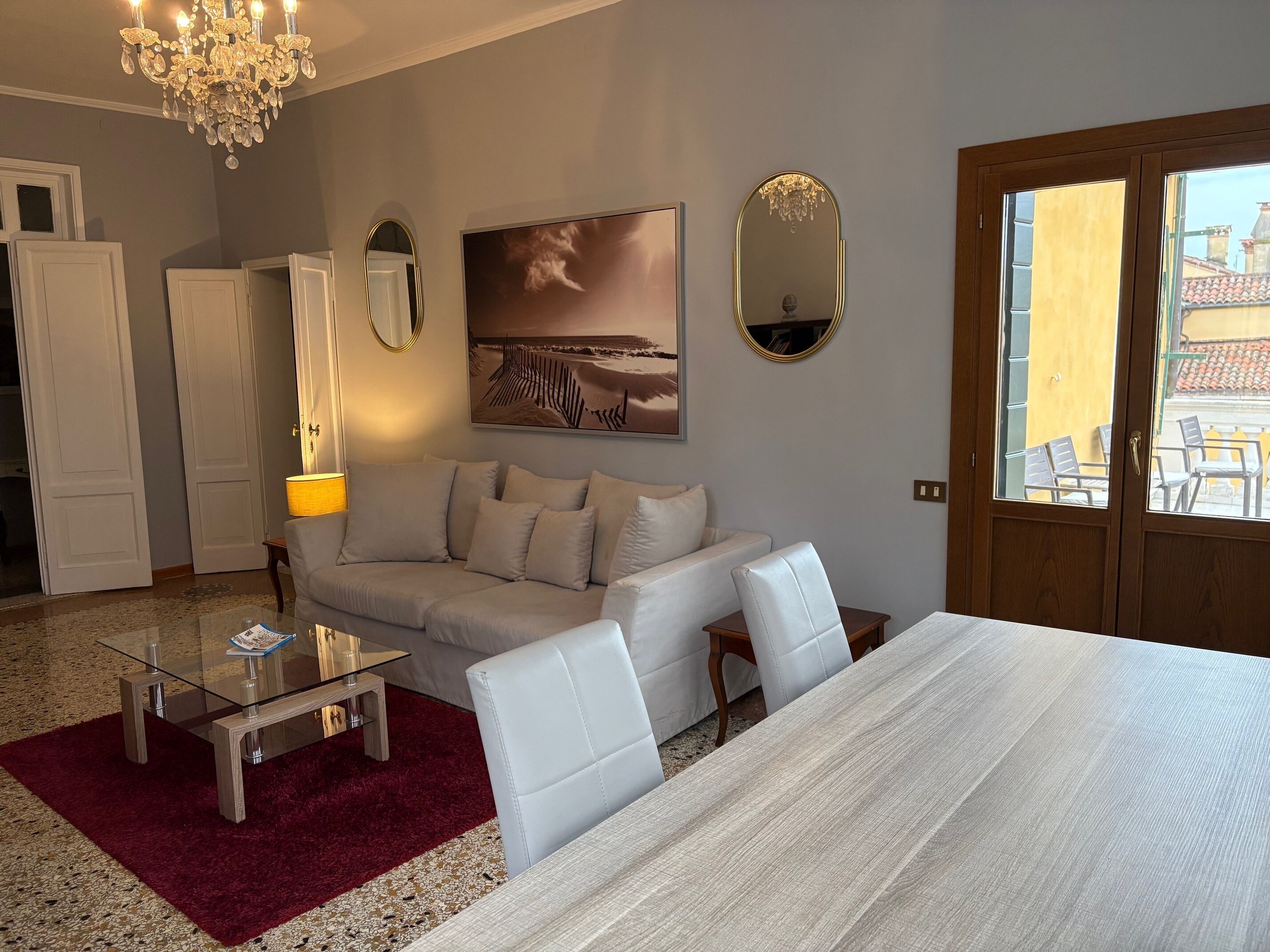 A room with a sofa, a glass coffee table, a wooden dining table, a red rug, a chandelier, a painting, and a view through a door.