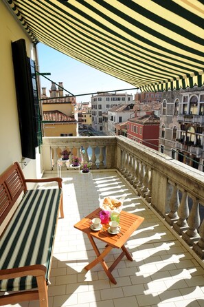 A balcony with a striped awning, a wooden bench, a small table with a cup and saucer, and potted plants.