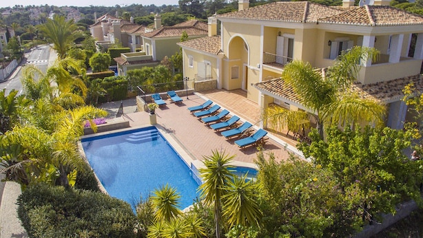 Ein Pool mit Liegestühlen, umgeben von üppiger Bepflanzung und einem Gebäude mit einem Ziegeldach.