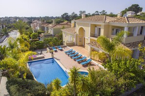 A pool with sun loungers, surrounded by lush greenery and a building with a tiled roof.