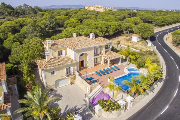 A two-story house with a swimming pool, surrounded by greenery and a curved road.