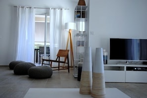 Un soggiorno moderno con televisore piatto, una sedia in legno, poggiapiedi e un mobiletto bianco.