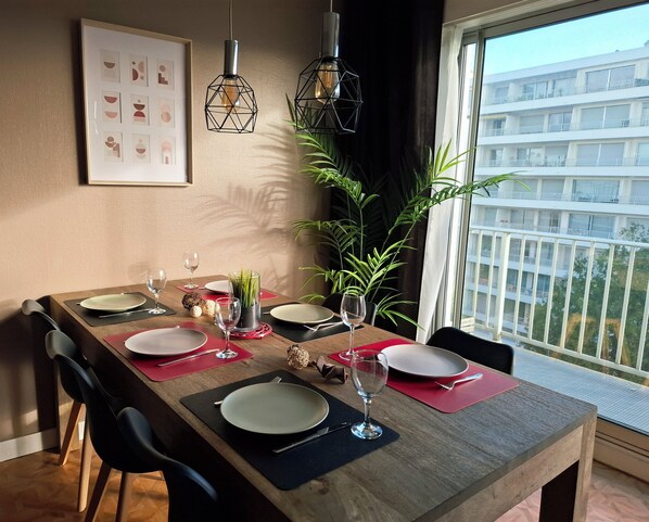 Un coin repas avec une table en bois dressée pour quatre personnes, une plante en pot et une vue sur un bâtiment par la fenêtre.