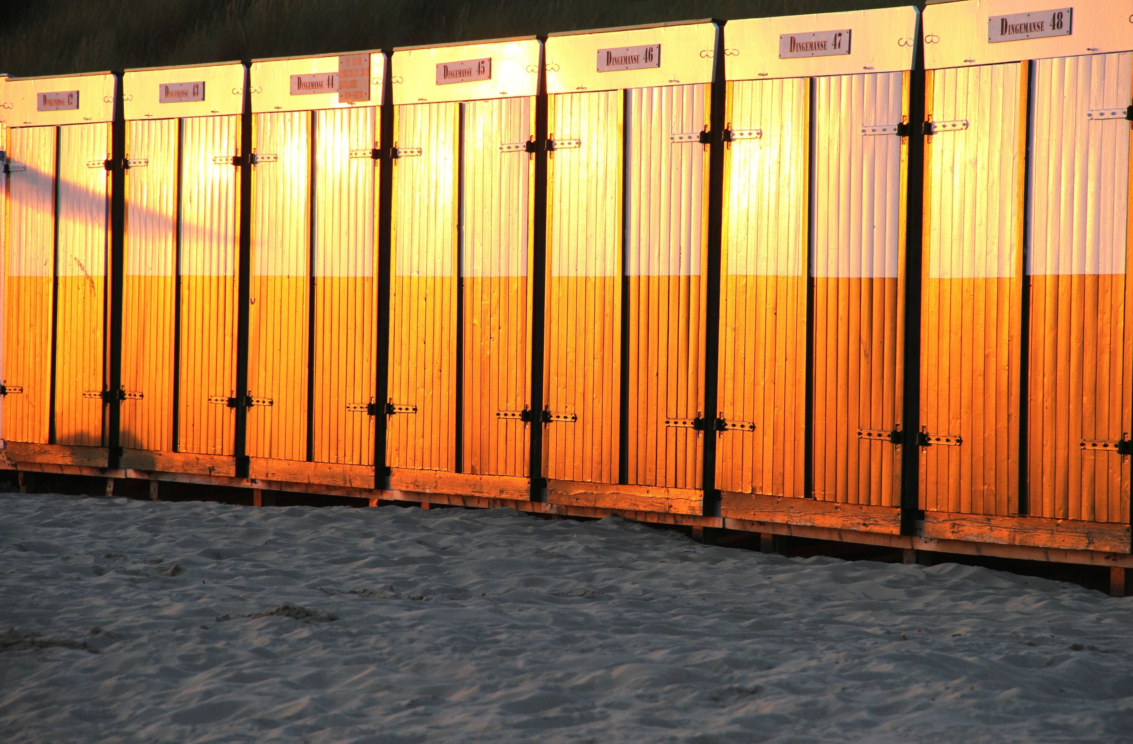 A row of wooden lockers with metal latches, labeled with numbers, on a sandy surface.