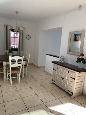 A dining area with a wooden table and white chairs, a white cabinet with drawers, and a window with curtains. - Villa in Puerto del Carmen, Lanzarote, Canary Islands (Tías)