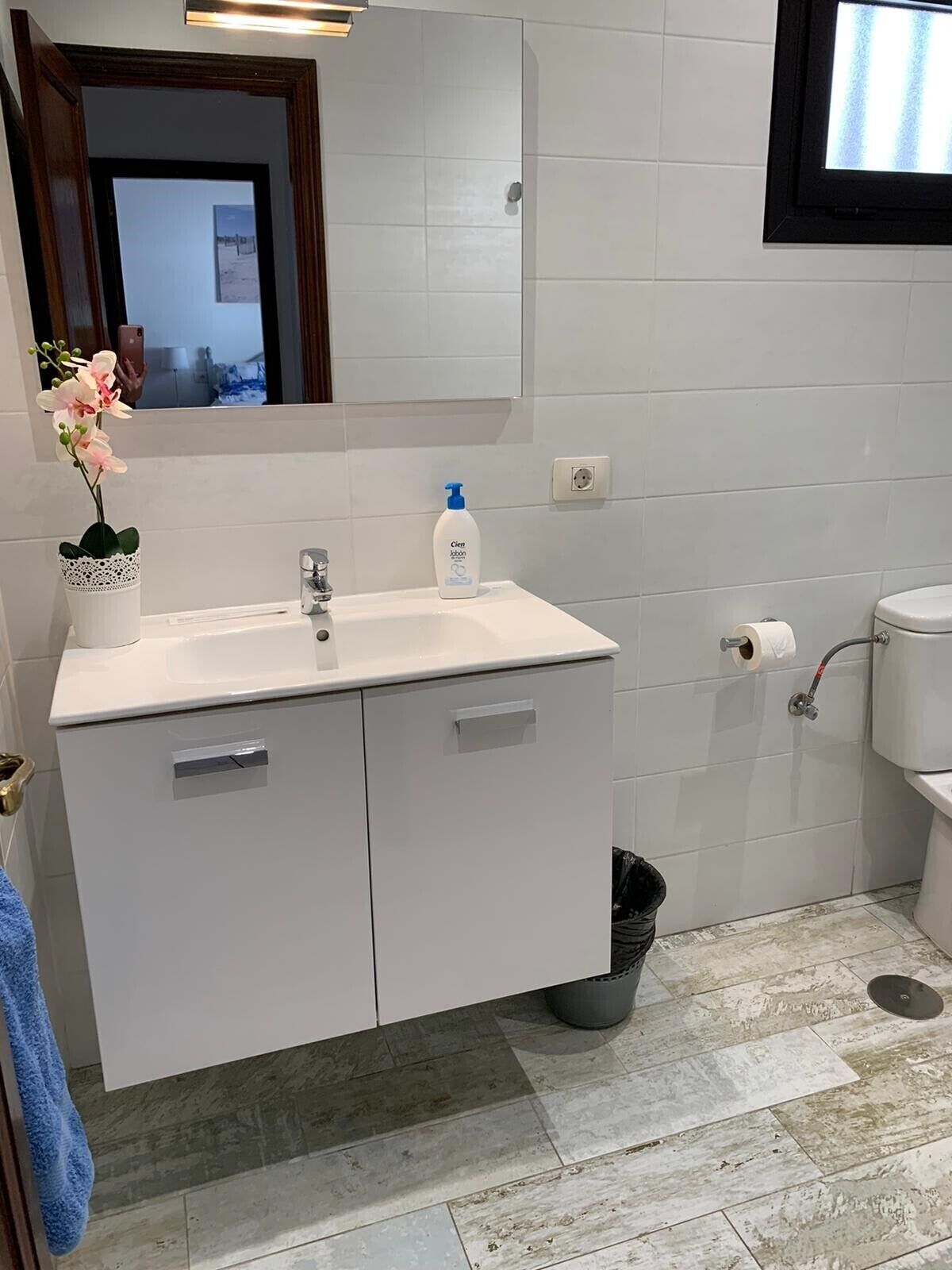 A bathroom with a white sink, mirror, and toilet. There is a trash bin and a blue towel hanging on the wall.