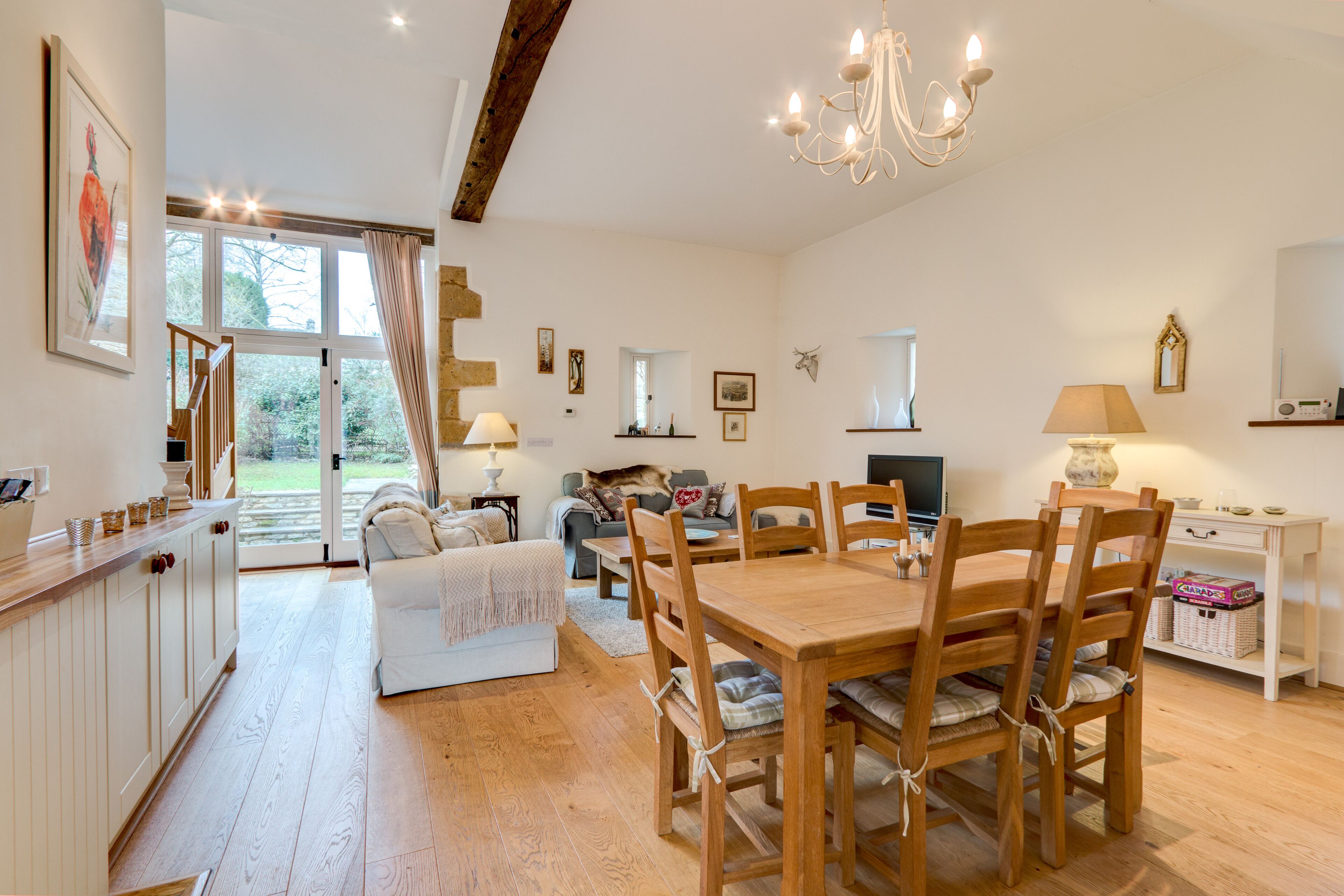 A spacious living area with a wooden dining table, chairs, a sofa, and a television. There is a kitchen area with cabinets and a countertop.