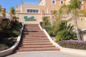 A hotel entrance with a sign reading "Beach Club Golf Resort," a set of stairs leading up to the entrance, and surrounding tropical landscaping. - 3 bedroomed villa with a south facing private garden access to golf and pool (Lagos)