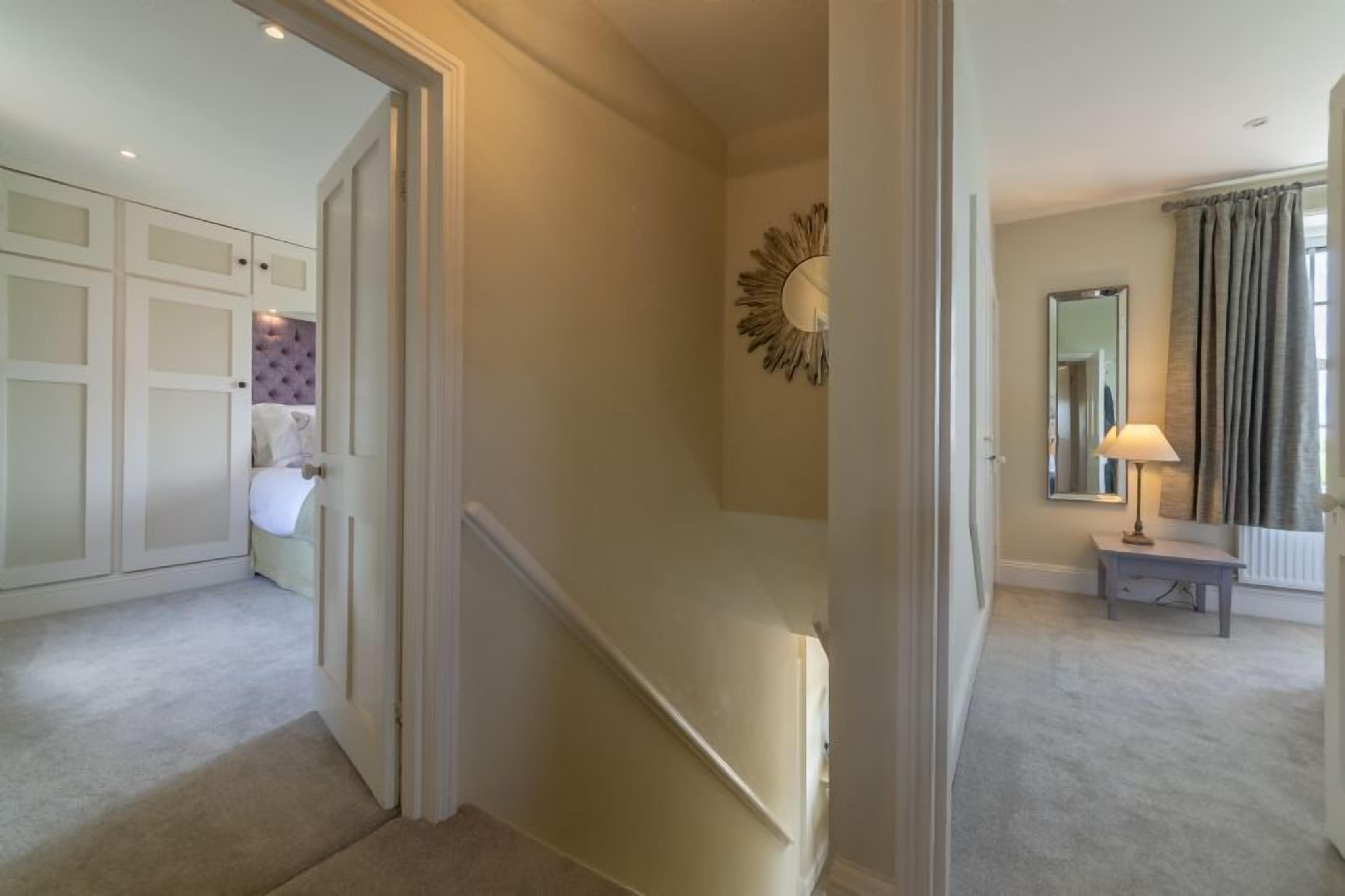 A hallway with a staircase, a door leading to a room with a bed, and a small table with a lamp.