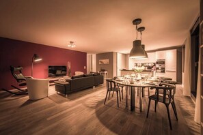 A modern living room with a sofa, dining table, and chairs, a TV, and a kitchen in the background.
