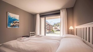 Ein Hotelzimmer mit einem großen Bett, einem Schreibtisch, einem Sessel, einem Gemälde und Blick auf Gebäude und Berge.