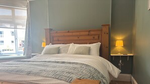 A neatly made bed with a wooden headboard, a bedside table with a lamp, and a view of the outside through a window. - St Matthias Lodge - Delightful 2 bed cottage  (Torquay)